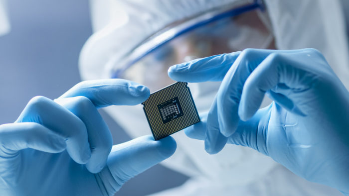 In Ultra Modern Electronic Manufacturing Factory Design Engineer in Sterile Coverall Holds Microchip with Gloves and Examines it.